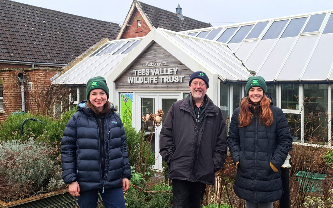 Three people stand outside a building. They are wearing warm outdoor clothes and hats with the Open Country logo on. The building has a sign which says Tees Valley Wildlife Trust.